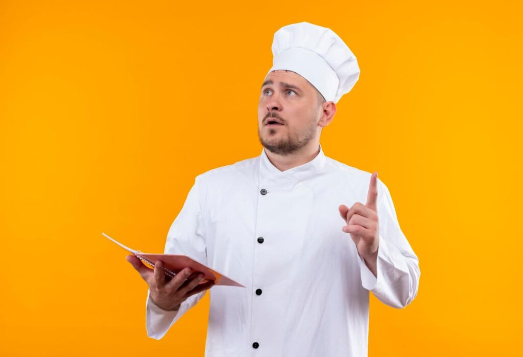 Chef in uniform holding notepad and raising finger while looking up on orange wall
