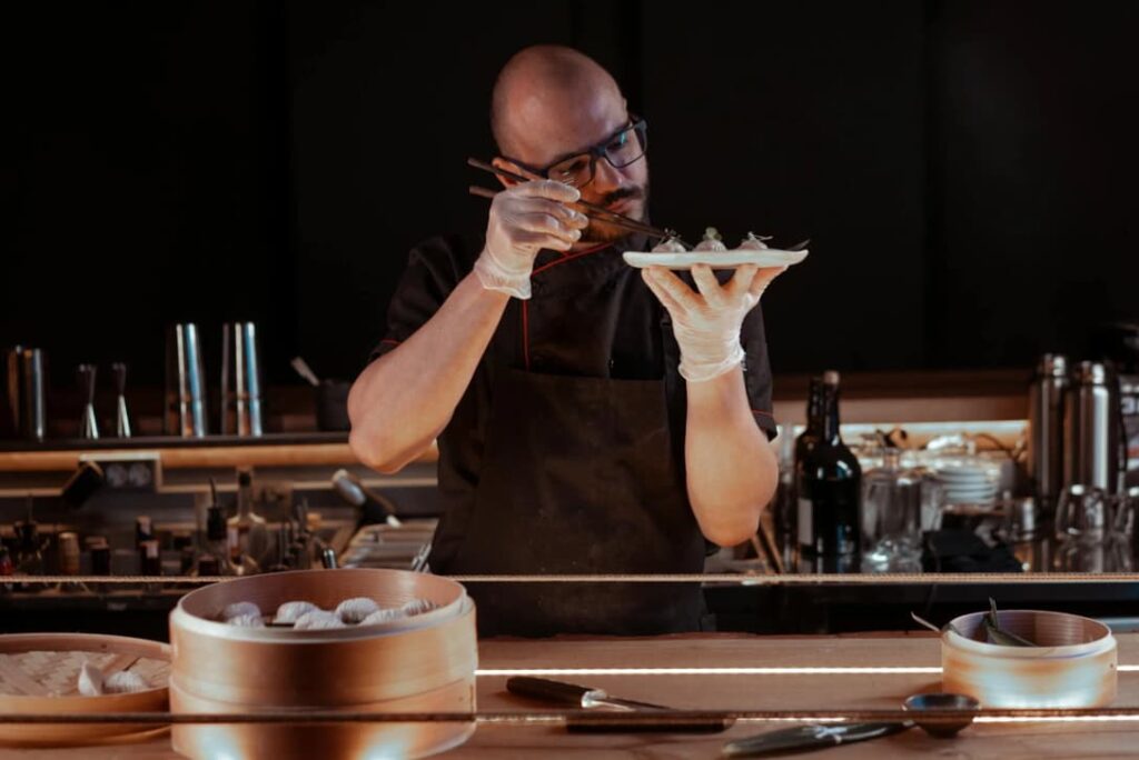 A chef is carefully garnishing a dish with tweezers in a dimly lit kitchen