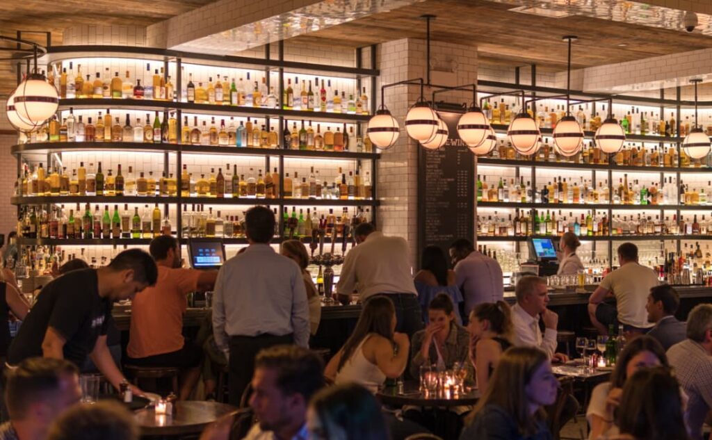 A bustling bar scene with a backlit wall of assorted bottles and a crowded, lively atmosphere