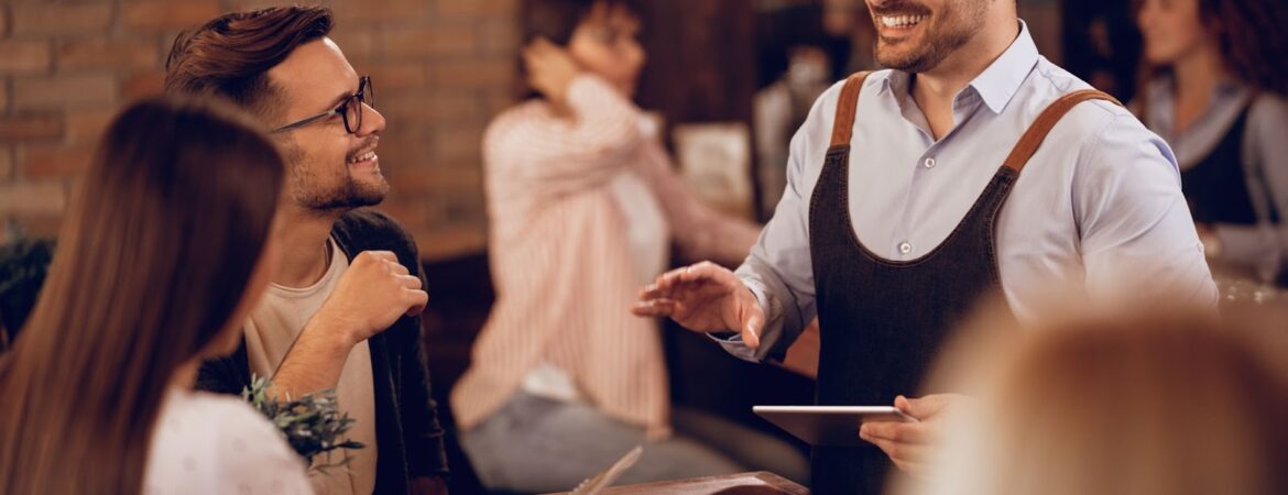 Smiling waiter holding digital tablet while taking order from a couple in a bar